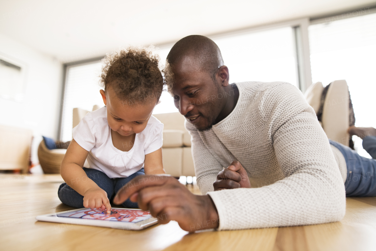 Young afro-american father at home with his cute little daughter lying on the floor watching something or playing a game on tablet.