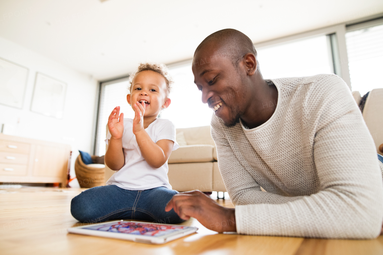 Young afro-american father at home with his cute little daughter lying on the floor watching something or playing a game on tablet.