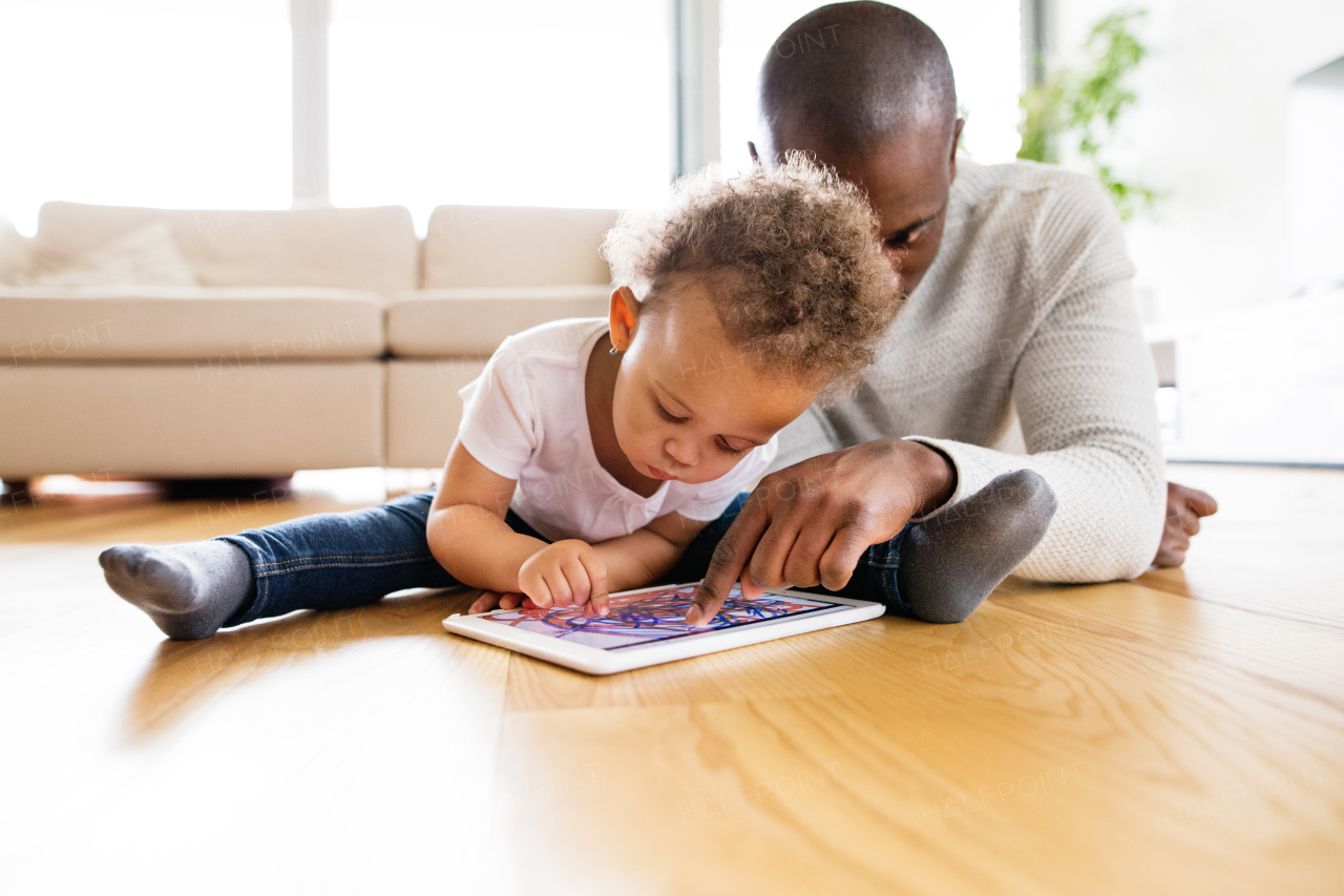 Young afro-american father at home with his cute little daughter lying on the floor watching something or playing a game on tablet.