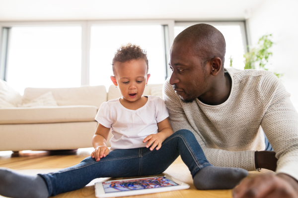 Young afro-american father at home with his cute little daughter lying on the floor watching something or playing a game on tablet.