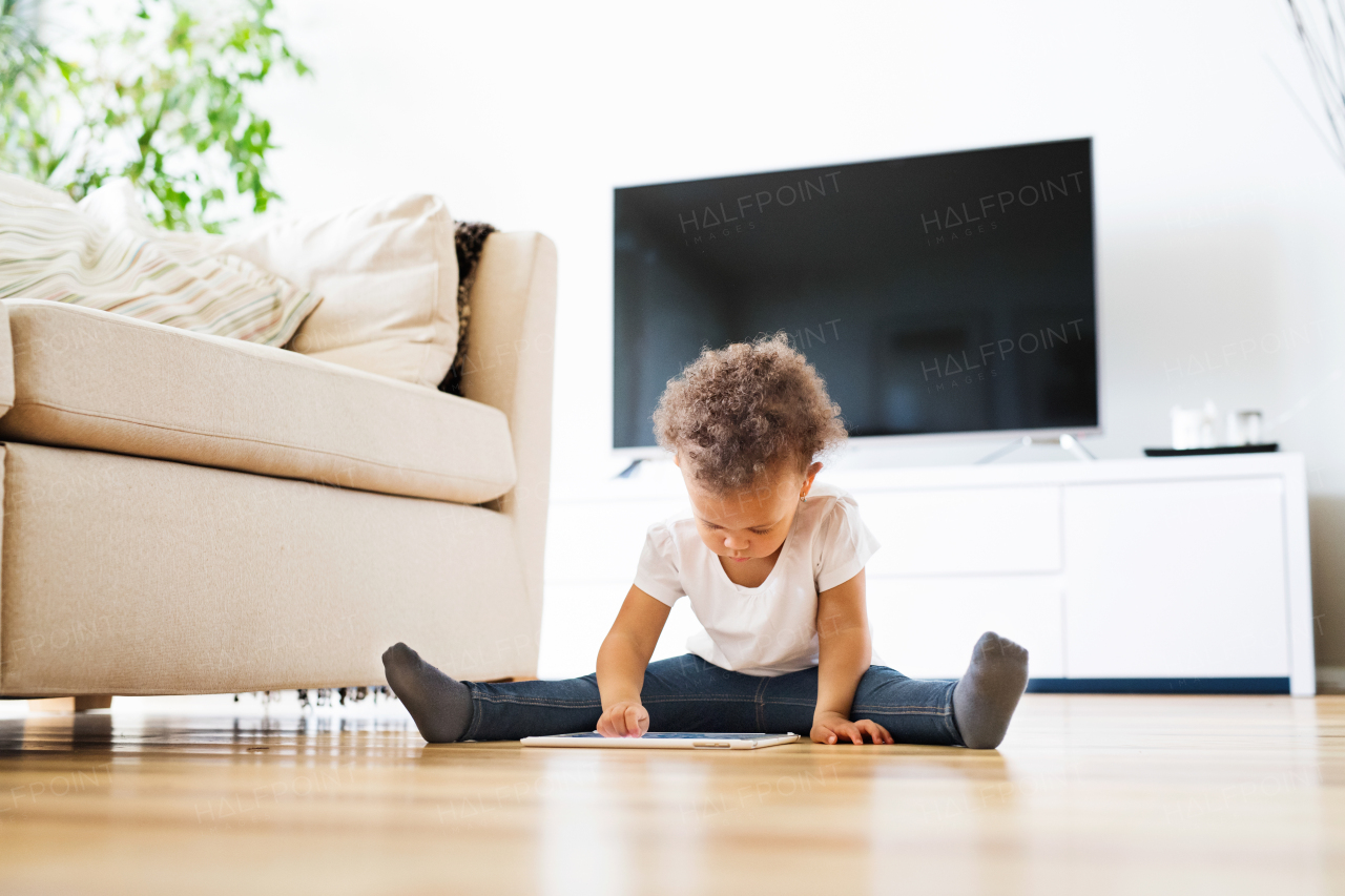 Cute little mixed-race girl with curly hair at home sitting on wooden floor playing game on tablet.