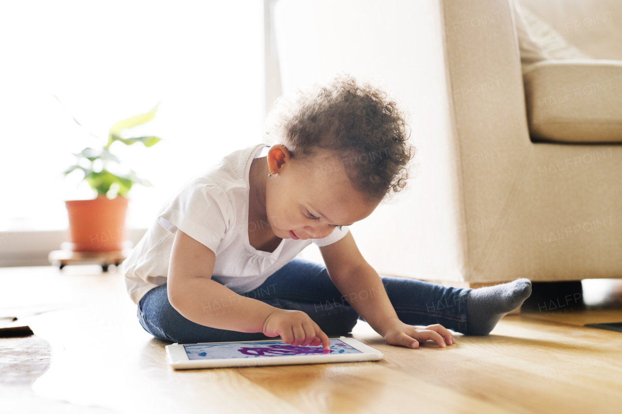 Cute little mixed-race girl with curly hair at home sitting on wooden floor playing game on tablet.