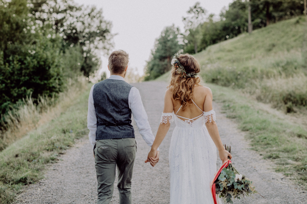 Beautiful young bride and groom outside in green nature holding hands. Rear view.