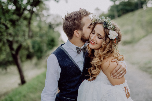 Beautiful young couple, groom and bride with flower wreath outside in green nature. A man kissing a woman.