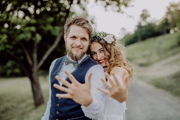 Beautiful young couple, groom and bride with flower wreath outside in green nature, showing wedding rings