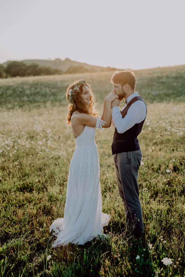 Beautiful young bride and groom outside in green nature at romantic sunset. Man kissing womans hands.