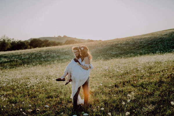 Beautiful young bride and groom outside in green nature at romantic sunset. Groom carrying bride in his arms.