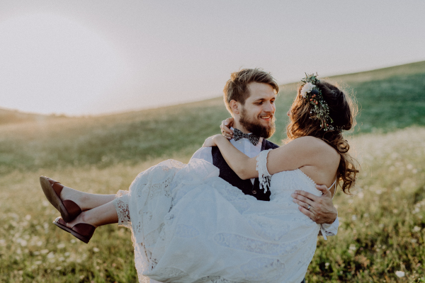 Beautiful young bride and groom outside in green nature at romantic sunset. Groom carrying bride in his arms.