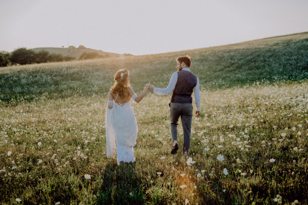 Beautiful young bride and groom outside in green nature at romantic sunset. Rear view.