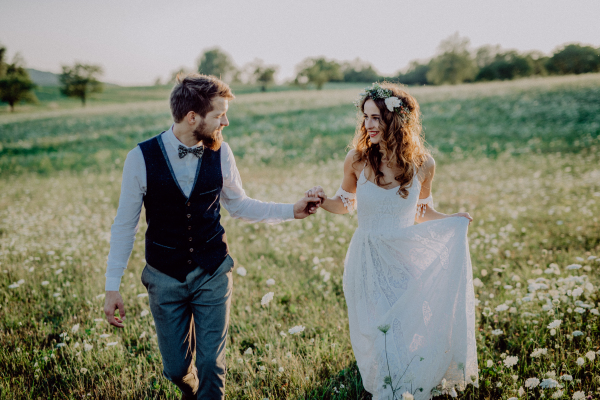 Beautiful young bride and groom outside in green nature at romantic sunset, holding hands.