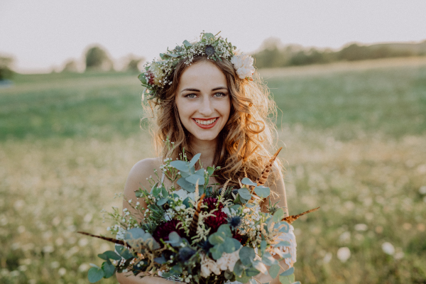 Beautiful young bride with flower wreath and a bouquet outside in green nature at sunset.