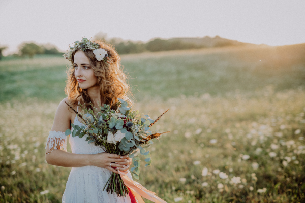 Beautiful young bride with flower wreath outside in green nature at sunset.