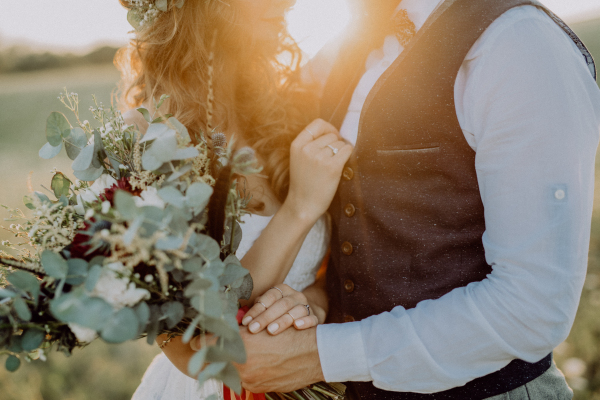 Unrecognizable young bride and groom outside in green nature at romantic sunset.