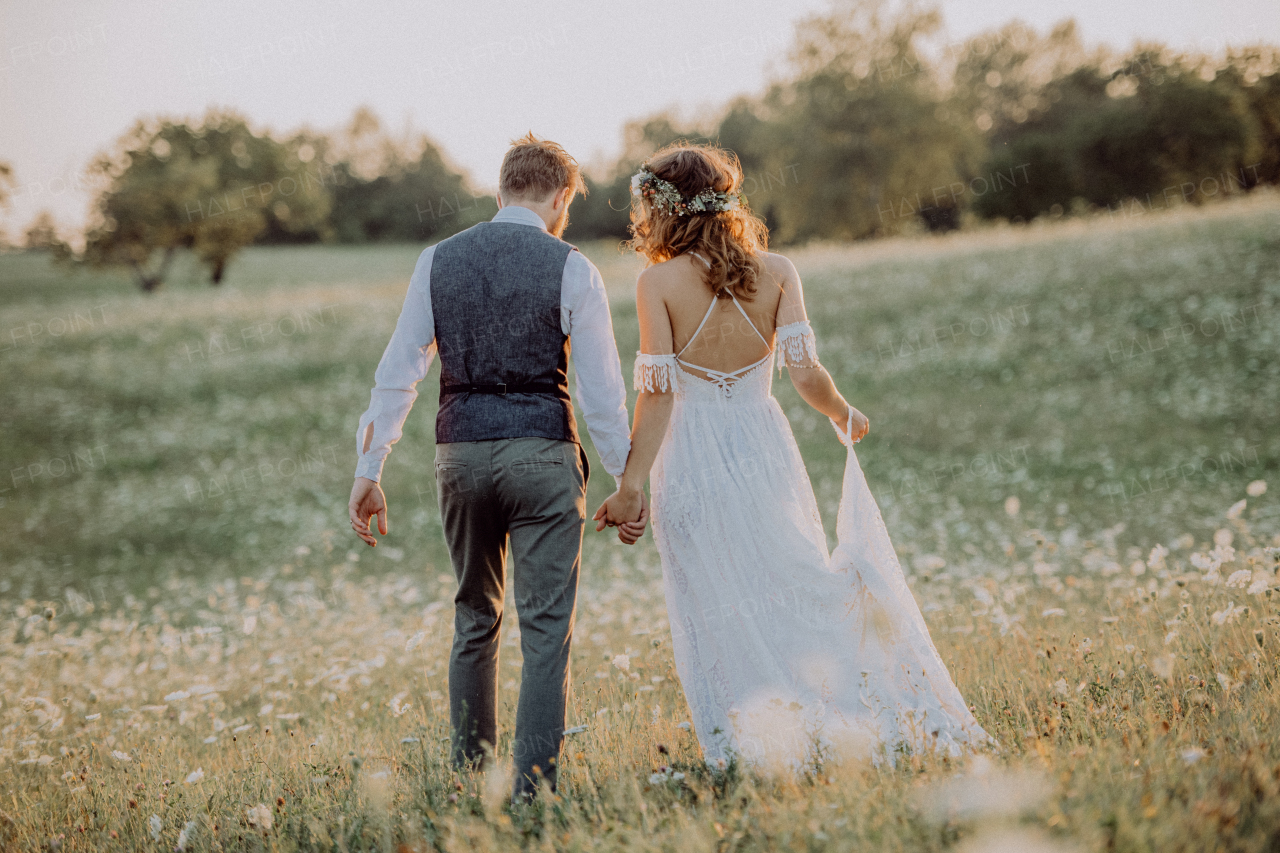 Beautiful young bride and groom outside in green nature at romantic sunset. Rear view.