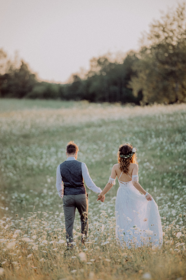 Beautiful young bride and groom outside in green nature at romantic sunset. Rear view.