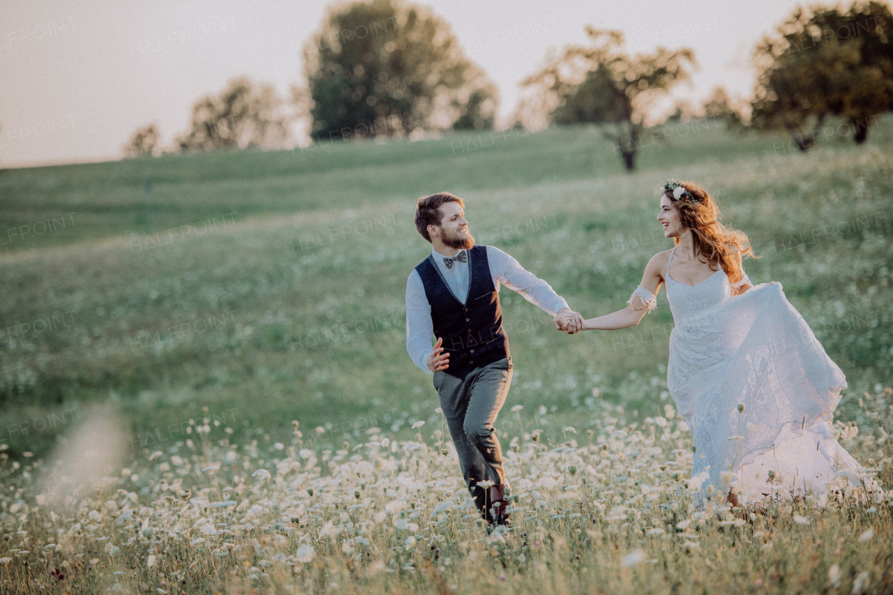 Beautiful young bride and groom outside in green nature at romantic sunset, holding hands.