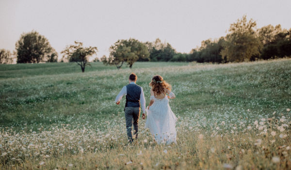 Beautiful young bride and groom outside in green nature at romantic sunset. Rear view.