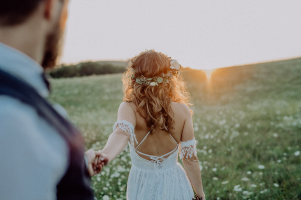 Beautiful young bride and groom outside in green nature at romantic sunset, holding hands. Rear view.