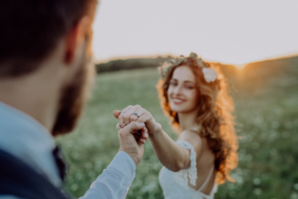 Beautiful young bride and groom outside in green nature at romantic sunset, holding hands.