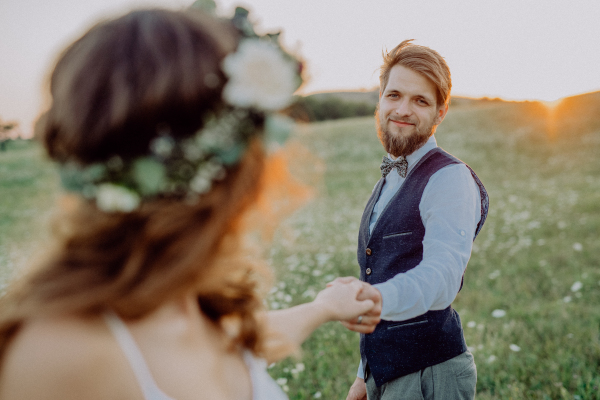 Beautiful young bride and groom outside in green nature at romantic sunset, holding hands.