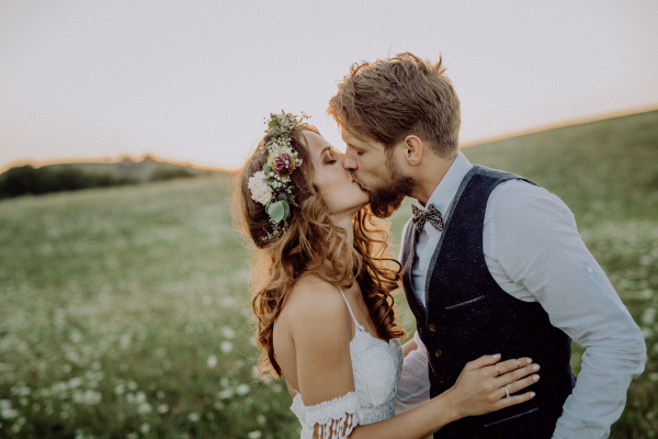 Beautiful young bride and groom kissing outside in green nature at romantic sunset.