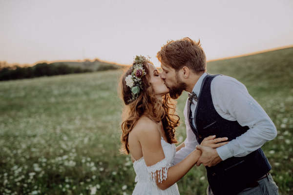 Beautiful young bride and groom kissing outside in green nature at romantic sunset.