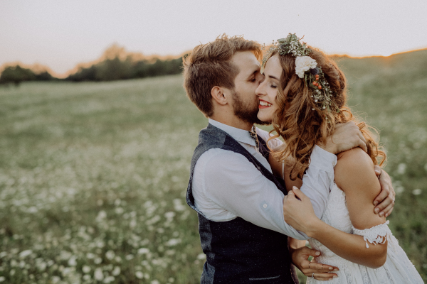 Beautiful young bride and groom kissing outside in green nature at romantic sunset.