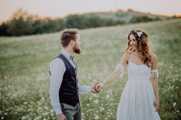 Beautiful young bride and groom outside in green nature at romantic sunset, holding hands.