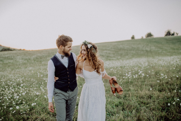 Beautiful young bride and groom outside in green nature at romantic sunset, walking. Bride holding shoes in her hand.