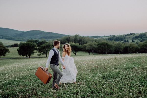 Beautiful young bride and groom outside in green nature at romantic sunset, holding hands.