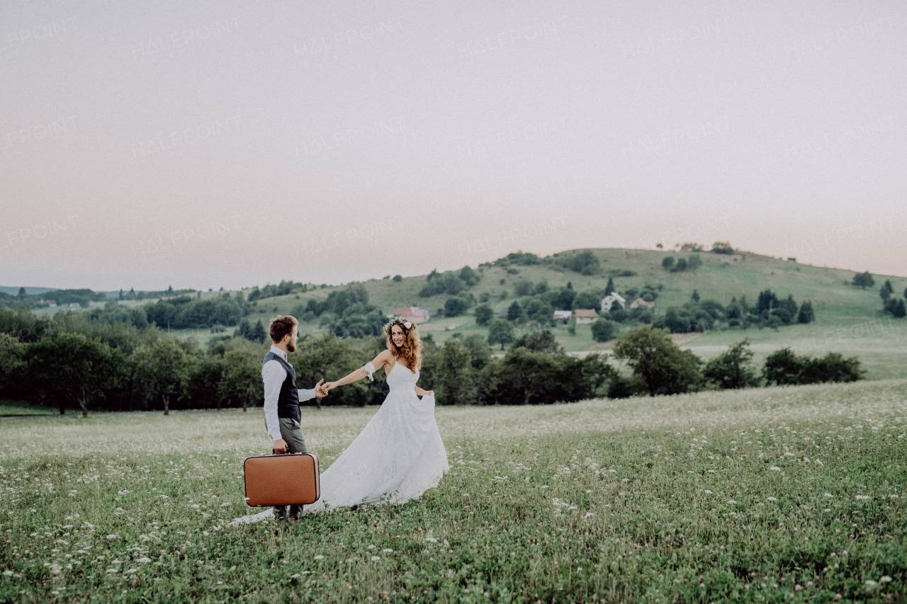 Beautiful young bride and groom outside in green nature at romantic sunset, holding hands.