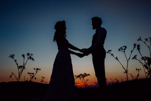 Silouethes of bride and groom holding hands and looking at each other on a meadow at night.