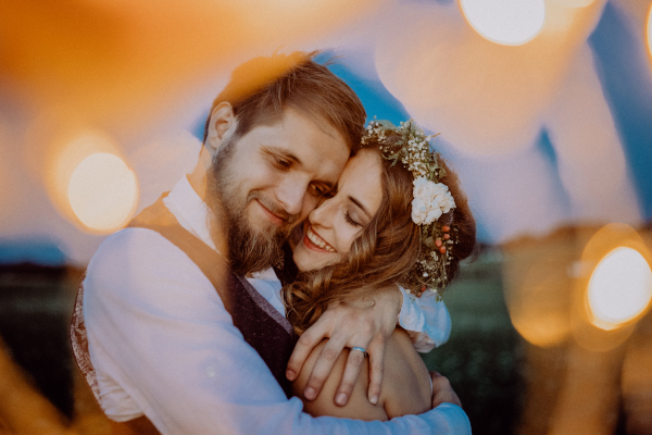 Beautiful young bride and groom at night on a meadow surrounded by chain of lights, hugging.