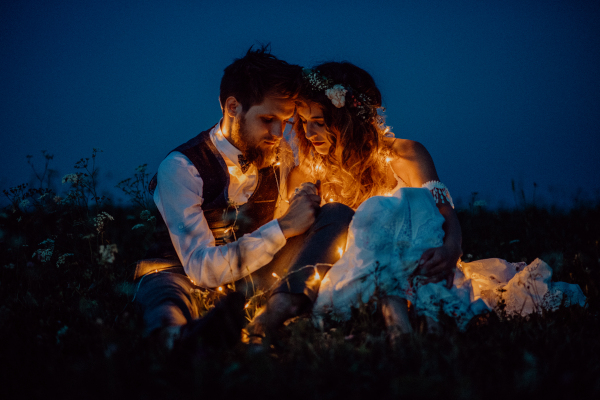 Beautiful young bride and groom at night on a meadow surrounded by chain of lights.