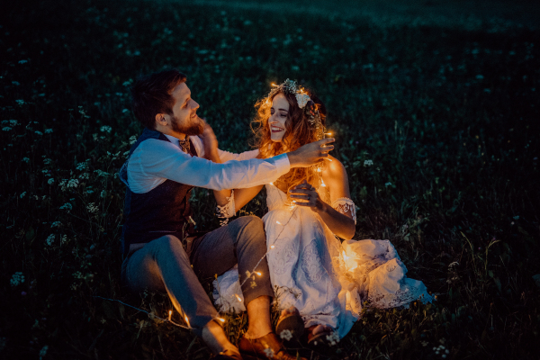 Beautiful young bride and groom at night on a meadow surrounded by chain of lights.