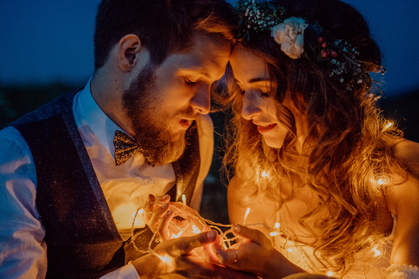 Beautiful young bride and groom at night on a meadow surrounded by chain of lights.