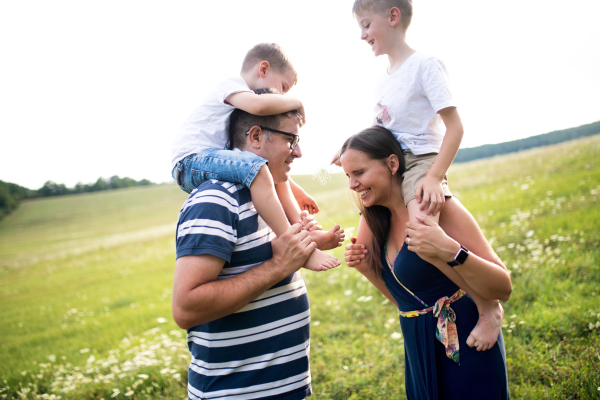 A young father and mother giving piggyback ride to small sons in nature on a summer day, laughing.