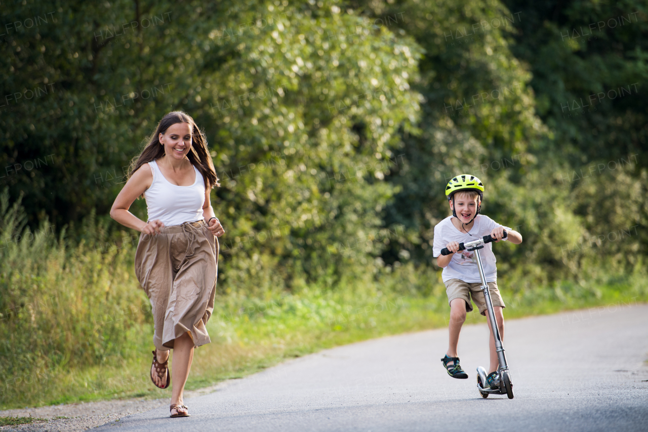 A cheerful small boy riding scooter and mother running on a road in park on a summer day.