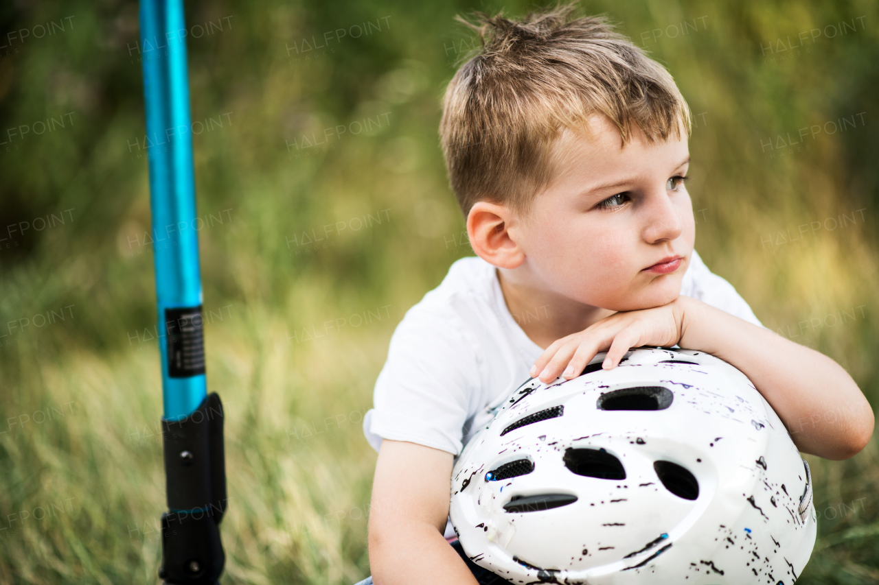 A thoughtful small boy with a helmet sitting on scooter on a road in park on a summer day.