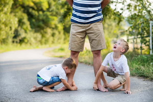 Two small sons playing with unrecognizable father on a road in park on a summer day, drawing with chalk.