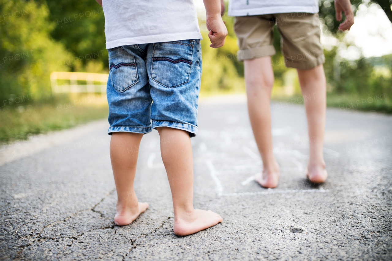 A rear view of legs of two barefoot unrecognizable small boys hopscotching on a road in park on a summer day.