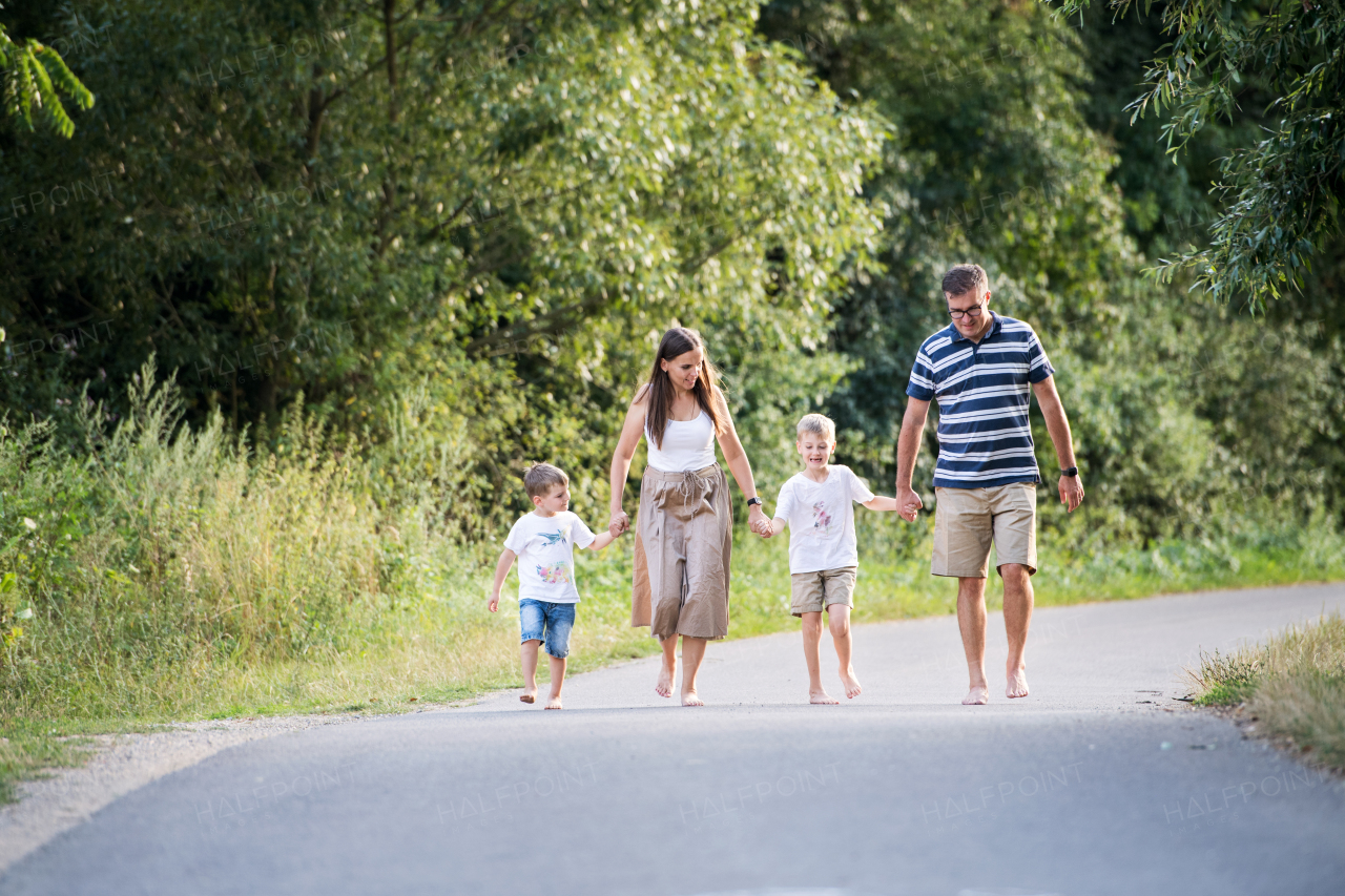 A young family with two small cheerful sons walking barefoot on a road in park on a summer day, holding hands.