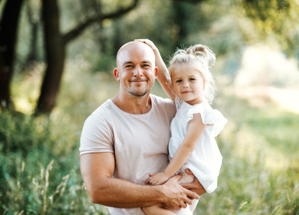 Handsome young father in green sunny summer nature holding his cute small daughter in the arms.