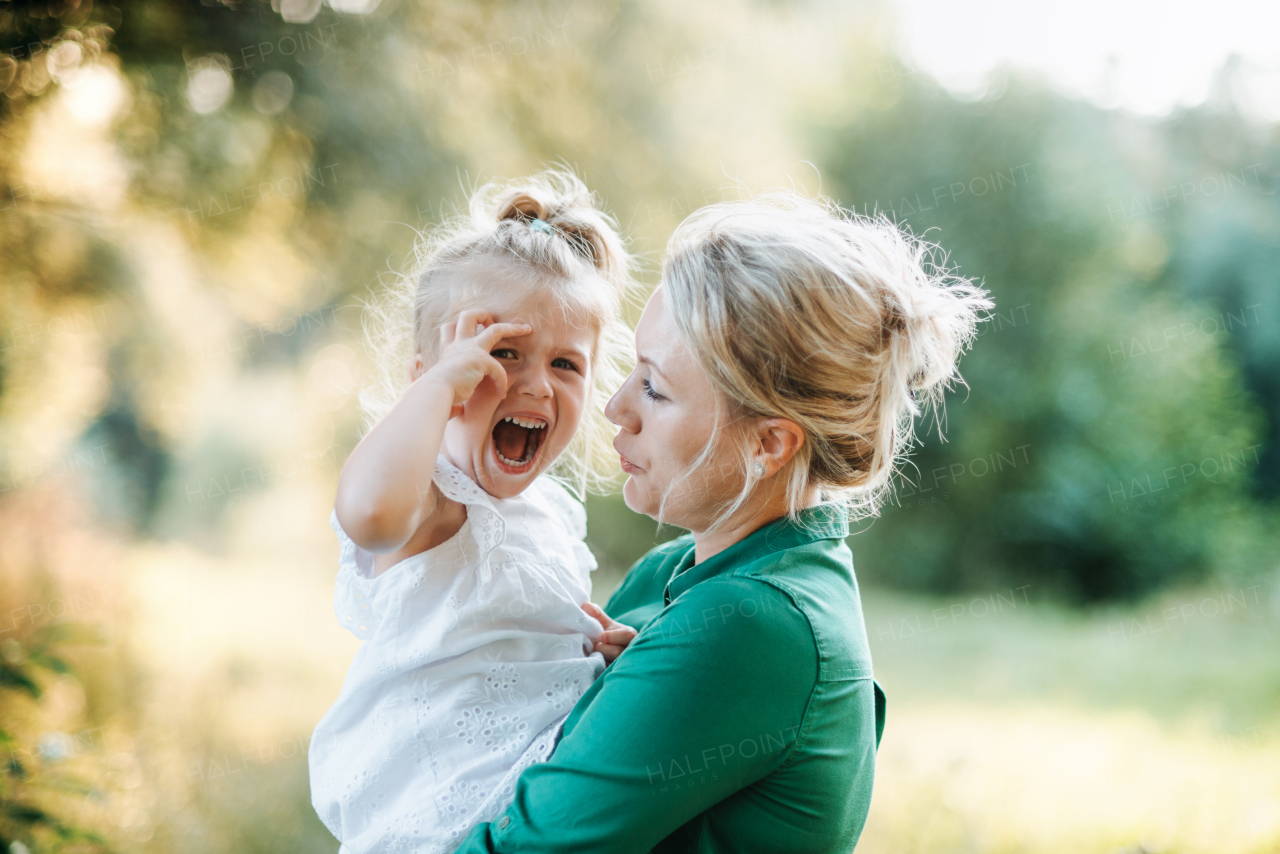 Young mother comforting crying small daughter in summer in nature, holding her.