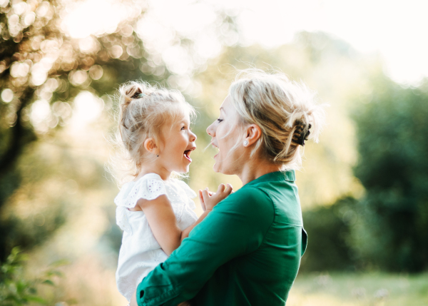 Beautiful young mother in green sunny summer nature holding her cute small daughter in the arms.