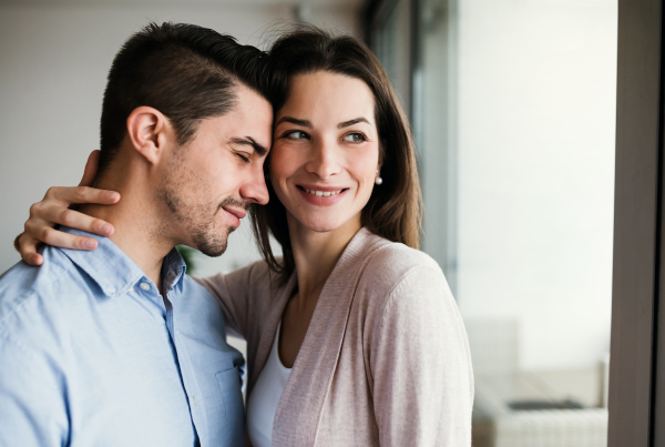 Young happy couple in love standing by the window at home, hugging. Copy space.