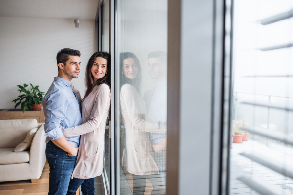 Young happy couple in love standing by the window at home, hugging. Copy space.