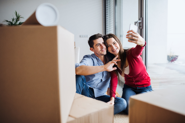 Young couple with cardboard boxes and smartphone moving in a new home, taking selfie.