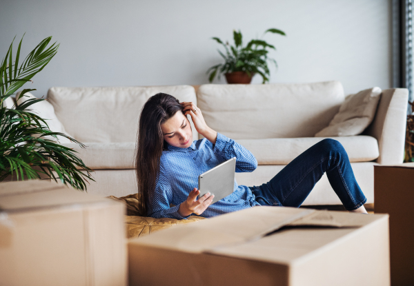 A young woman with tablet and cardboard boxes lying on the floor, moving in a new home.