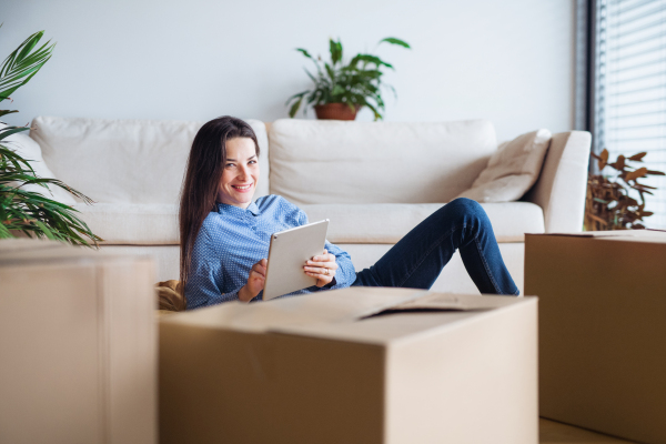 A young woman with tablet and cardboard boxes lying on the floor, moving in a new home.